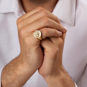 May include: A gold signet ring with an engraved design. The ring is round with a detailed emblem in the center. The ring is worn on a finger. The person is wearing a white dress shirt.
