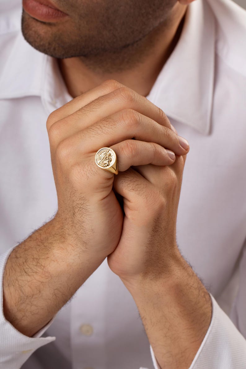 May include: A gold signet ring with an engraved design. The ring is round with a detailed emblem in the center. The ring is worn on a finger. The person is wearing a white dress shirt.
