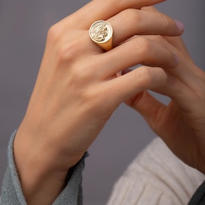 May include: A gold signet ring with a detailed circular emblem. The ring is worn on a finger, showcasing its polished surface and intricate design. The ring's design features a detailed relief.