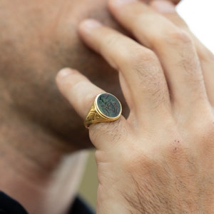 Peut inclure: Une bague signet en or avec une pierre ovale verte orn&eacute;e d'un motif grav&eacute; d&eacute;taill&eacute;. La bague est port&eacute;e &agrave; l'index d'une main.