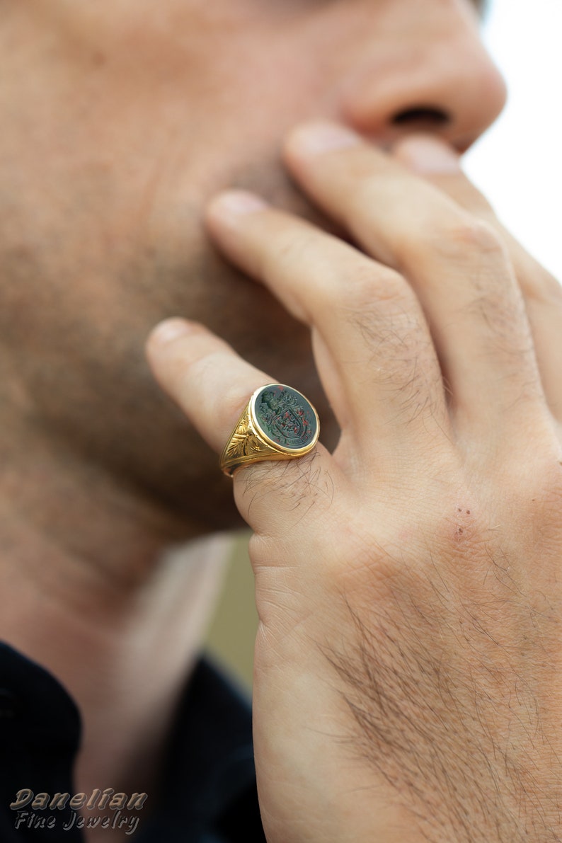 Peut inclure: Une bague signet en or avec une pierre ovale verte orn&eacute;e d'un motif grav&eacute; d&eacute;taill&eacute;. La bague est port&eacute;e &agrave; l'index d'une main.