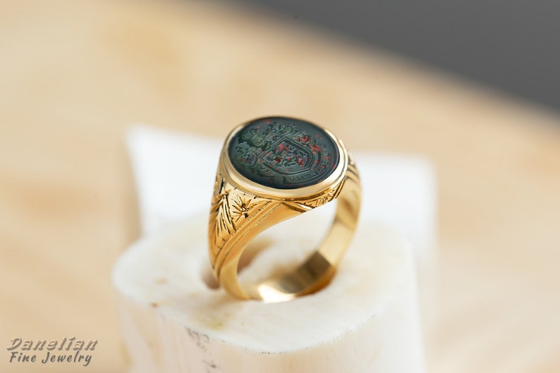 Peut inclure: Une bague signet en or avec un cam&eacute;e vert et rouge repr&eacute;sentant un blason. La bague est orn&eacute;e d'un motif floral d&eacute;taill&eacute; sur la bande.