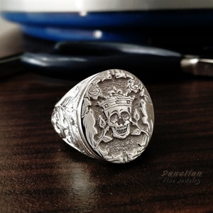 Puede incluir: Un anillo de sello de plata con un grabado detallado de una calavera con corona, rodeada de enredaderas de rosas. La banda tiene intrincados patrones de volutas en los lados. El texto Danelian Fine Jewelry es visible en la esquina inferior derecha.