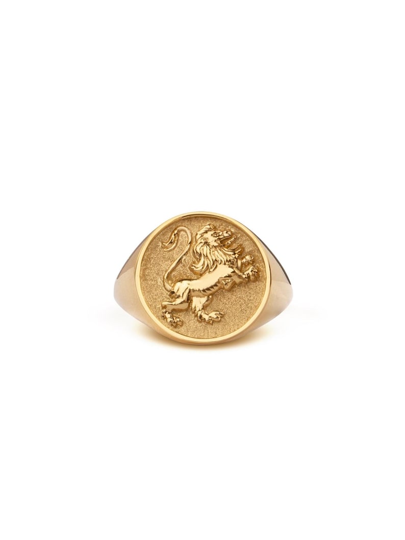 Puede incluir: Anillo de sello dorado con un relieve detallado de un le&oacute;n. El anillo tiene una cara redondeada con un fondo texturizado y una banda pulida. El le&oacute;n est&aacute; representado en una pose din&aacute;mica, con su melena y cola fluyendo.
