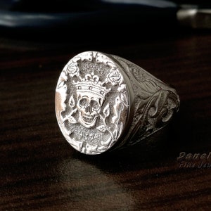 Puede incluir: Un anillo de sello de plata con un dise&ntilde;o central en relieve de una calavera con corona, flanqueado por motivos florales sobre un fondo texturizado. La banda muestra intrincados grabados de volutas. El texto Danelian Fine Jewelry es visible.