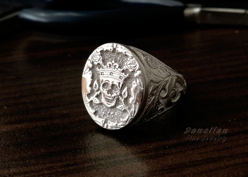 Puede incluir: Un anillo de sello de plata con un dise&ntilde;o central en relieve de una calavera con corona, flanqueado por motivos florales sobre un fondo texturizado. La banda muestra intrincados grabados de volutas. El texto Danelian Fine Jewelry es visible.