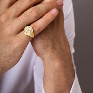 Puede incluir: Un anillo de sello dorado con un emblema detallado de una criatura alada. El anillo se lleva en un dedo, sobre una camisa blanca y un fondo neutro. El dise&ntilde;o del anillo es el punto focal, mostrando su artesan&iacute;a.