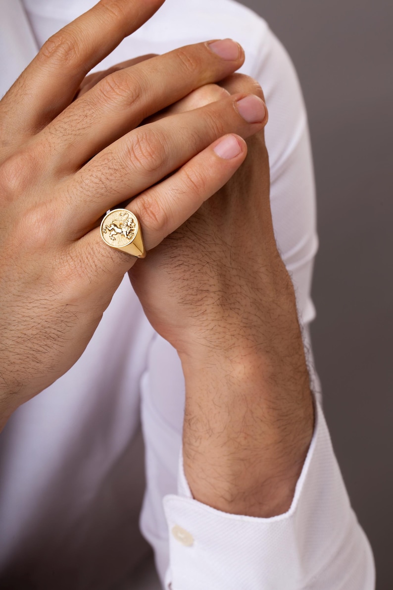 Puede incluir: Un anillo de sello dorado con un emblema detallado de una criatura alada. El anillo se lleva en un dedo, sobre una camisa blanca y un fondo neutro. El dise&ntilde;o del anillo es el punto focal, mostrando su artesan&iacute;a.