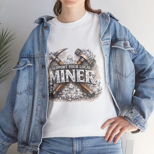 May include: White t-shirt with a graphic featuring a pickaxe and a hammer, along with the text "SUPPORT YOUR LOCAL MINER". The design includes crystals and a weathered aesthetic. The shirt is worn with a blue denim jacket.