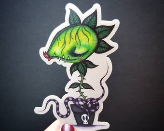 Puede incluir: Una planta de dibujos animados verde y morada con un ojo cerrado y una boca con dientes. La planta está en una maceta negra con un diseño de calavera. La planta está envuelta en una serpiente morada y blanca.