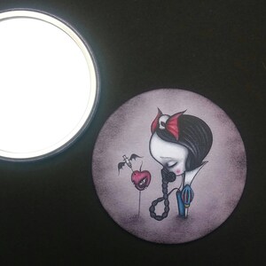 May include: A round, compact mirror with a dark gray background featuring a stylized illustration of Snow White with a red apple and a syringe. The illustration is in a dark, gothic style.