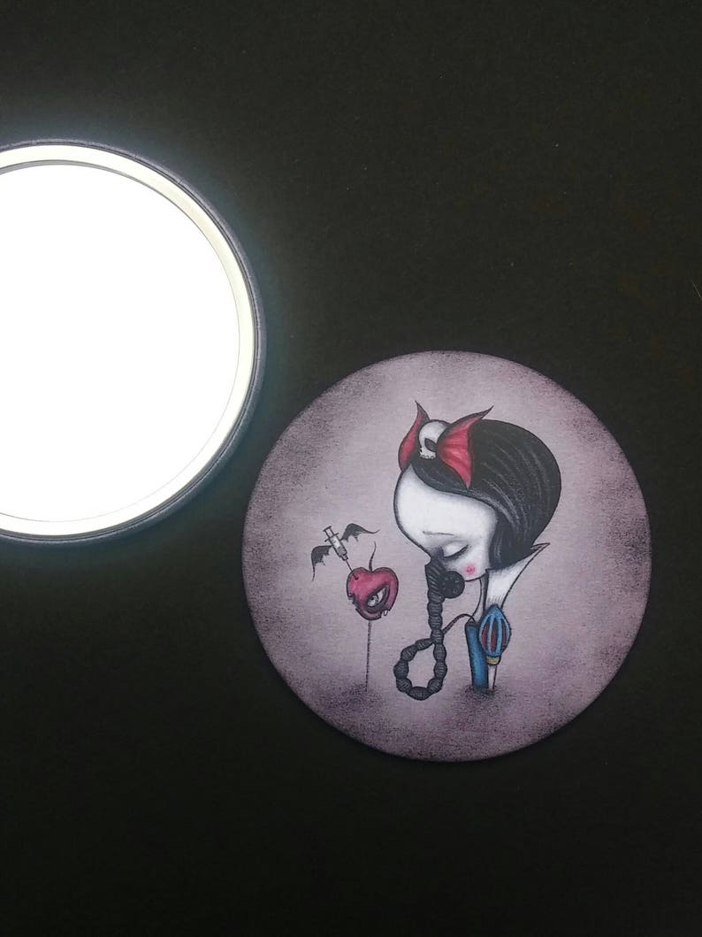 May include: A round, compact mirror with a dark gray background featuring a stylized illustration of Snow White with a red apple and a syringe. The illustration is in a dark, gothic style.