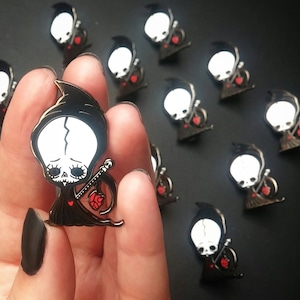 Puede incluir: Una colección de pines de esmalte negro con una figura de esqueleto de dibujos animados con un corazón rojo. El esqueleto lleva una túnica negra con capucha y sostiene una guadaña. Los pines están dispuestos sobre una superficie negra.