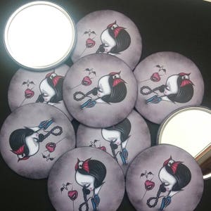 May include: Set of eight round compact mirrors featuring a dark-haired woman with a red headband and a red apple. The woman is depicted with a sad expression and a needle in her hand. The background is a dark gray color.