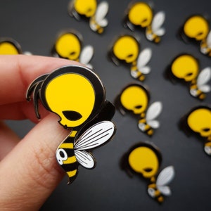 Peut inclure: Gros plan sur une épingle en émail noire et jaune en forme d'abeille avec un crâne pour tête. L'abeille est en position de vol, les ailes déployées. L'épingle est entourée d'autres épingles en forme d'abeille dans différentes positions.