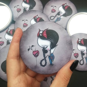 May include: A set of round compact mirrors featuring a dark-haired girl with a red headband and a red apple with a syringe. The girl is wearing a blue and white dress and a necklace with a noose. The background is a dark gray.