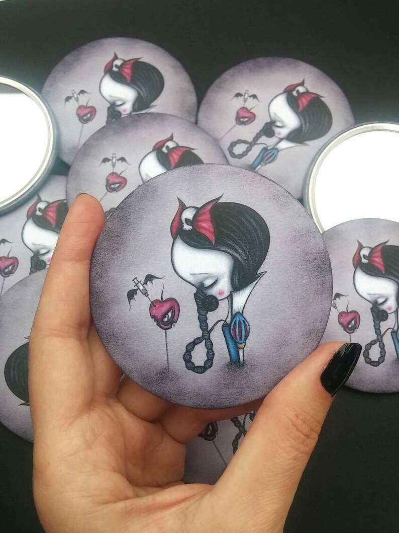 May include: A set of round compact mirrors featuring a dark-haired girl with a red headband and a red apple with a syringe. The girl is wearing a blue and white dress and a necklace with a noose. The background is a dark gray.