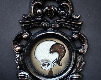 May include: A black ornate frame with a detailed design surrounds a whimsical illustration of a creature with a large head, brown hair, and a white face. The creature has a single large eye and a small mouth.