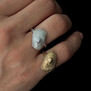 Op de afbeelding: Twee ringen met een gestructureerd, ovaal schelpontwerp. De ene ring is afgewerkt in een heldere zilverkleur, terwijl de andere is afgewerkt in een warme goudkleur. Beide ringen hebben een dunne, gladde band en een gedetailleerde, geribbelde textuur.