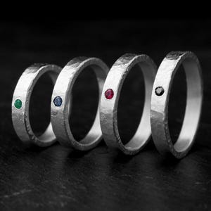 May include: Four silver rings with different colored gemstones. The rings are textured and have a hammered finish. The gemstones are emerald green, sapphire blue, ruby red, and black.