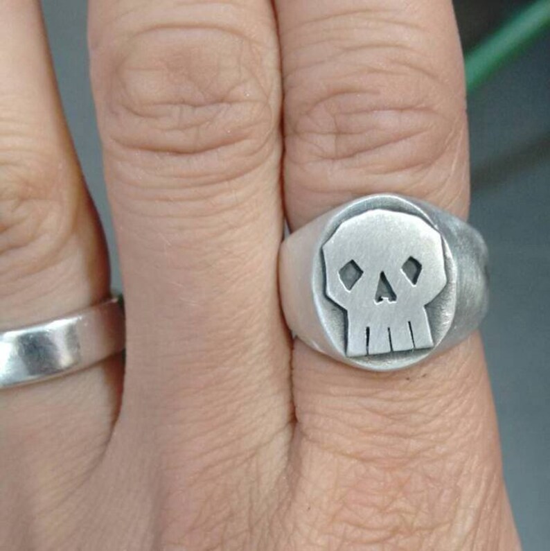 Pode incluir: Um anel de prata com um design de caveira. A caveira est&aacute; em um c&iacute;rculo e tem um design simples com duas &oacute;rbitas e um nariz.