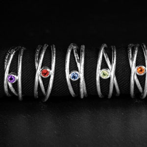 May include: Five silver rings with a textured finish, each featuring a different colored gemstone: purple, red, blue, green, and orange. The rings have a unique crisscross design, set against a black background.