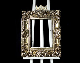 May include: A gold ornate picture frame with intricate details sits on a white easel against a black background.