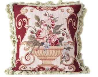 May include: A square throw pillow with a floral tapestry design. The pillow features a burgundy border with a cream-colored center. The center features a vase with pink and white roses and green leaves. The pillow has a green fringe border.