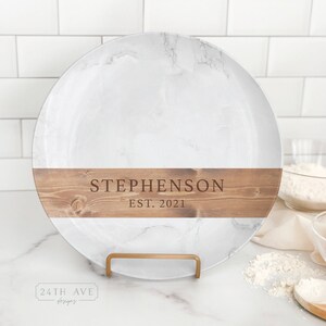 Puede incluir: Bandeja de servir de mármol blanco con una banda de madera marrón alrededor del borde. La banda de madera tiene el texto "STEPHENSON EST. 2021" en letras marrones. La bandeja está sobre un soporte de metal dorado.