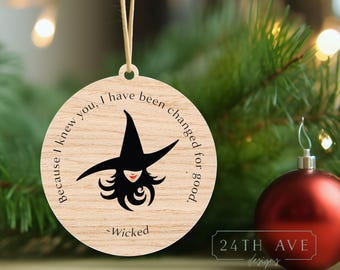 Puede incluir: Adorno navideño redondo de madera con una ilustración de bruja y el texto "Because I knew you, I have been changed for good. -Wicked." El adorno tiene una cuerda para colgar. Una bola roja en el fondo.