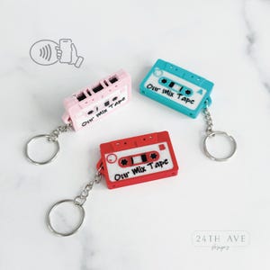 May include: Three colorful cassette tape keychains: pink, teal, and red. Each keychain has the text "Our Mix Tape" and a silver keyring. The keychains are on a white surface.