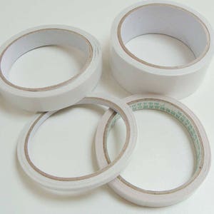 Peut inclure: Quatre rouleaux de ruban adhésif double face blanc. Le ruban est sur une surface blanche.