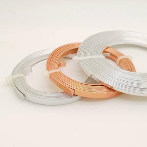 Puede incluir: Tres bobinas de alambre plano en colores plata, cobre y plata. Cada bobina se sujeta con una banda de plástico blanco. Los alambres están cuidadosamente enrollados, sobre fondo blanco.