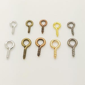 Peut inclure: Un ensemble de dix pitons à vis en métal disposés sur deux rangées. Les pièces présentent diverses finitions métalliques, notamment argent, laiton antique, cuivre, or et bronze à canon. Chaque élément se compose d'un anneau et d'une tige filetée.