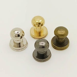 Può includere: Un set di quattro bottoni in metallo con una parte superiore arrotondata a pomello e una base circolare piatta. I bottoni presentano quattro distinte finiture metalliche: argento lucido, oro brillante, grigio canna di fucile e bronzo antico.