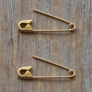 May include: Two gold-toned metal safety pins are positioned horizontally, one above the other. Each pin features a smooth, polished finish with a classic coiled spring mechanism at one end and a secure clasp closure at the other end.