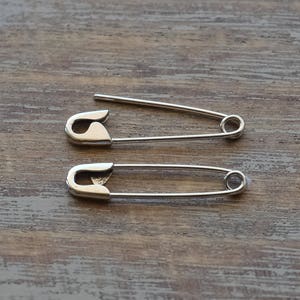 May include: Two silver-toned metal safety pins. The top safety pin is shown in an open position with the sharp needle point exposed, while the bottom safety pin is shown in a closed position with the needle secured inside the protective clasp.