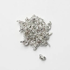 Op de afbeelding: Een cluster van kleine, onregelmatig gevormde, zilverkleurige metalen objecten. De objecten hebben een textuur, organisch uiterlijk, met een paar stukken los van de hoofdcluster. De achtergrond is effen wit.