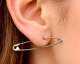 Könnte beinhalten: Ein silberner Sicherheitsnadel-Ohrring, der in einem durchbohrten Ohr getragen wird. Der Ohrring ist ein großes, stilisiertes Sicherheitsnadel-Design, bei dem der Verschluss der Nadel sichtbar ist. Ein schlichtes, modernes Accessoire.