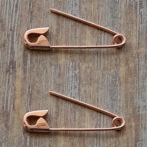 May include: Two rose gold-toned metal safety pins, each featuring a classic coil spring mechanism at one end and a secure clasp closure at the other. The pins are polished with a smooth, reflective surface and are positioned horizontally.
