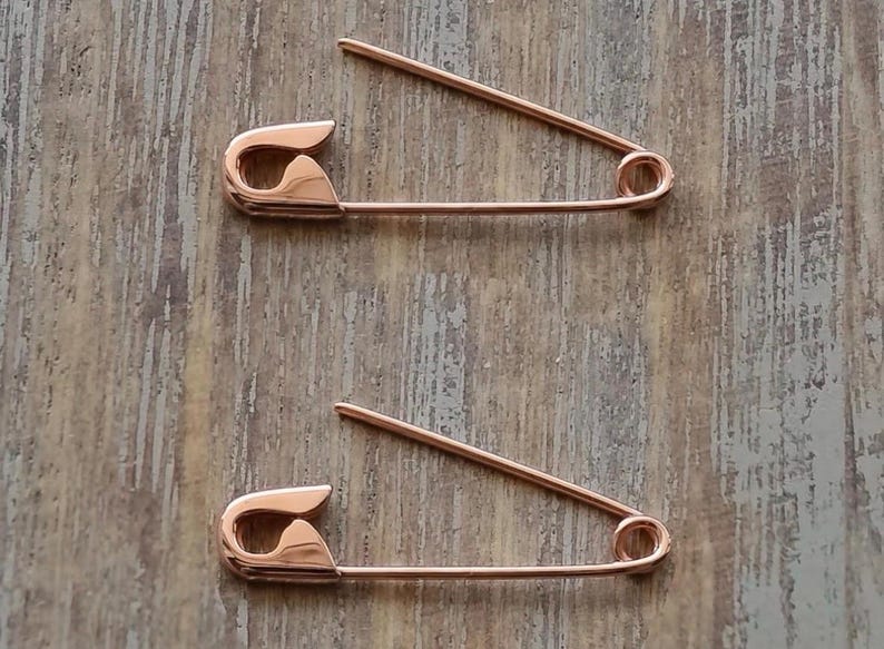 May include: Two rose gold-toned metal safety pins, each featuring a classic coil spring mechanism at one end and a secure clasp closure at the other. The pins are polished with a smooth, reflective surface and are positioned horizontally.