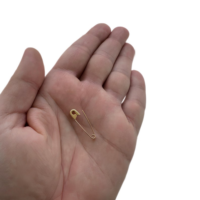 May include: A small, gold-toned safety pin featuring a classic coil spring mechanism at one end and a pointed tip that secures into a protective clasp. The metal surface has a smooth, polished finish, reflecting light across its slender, curved frame.