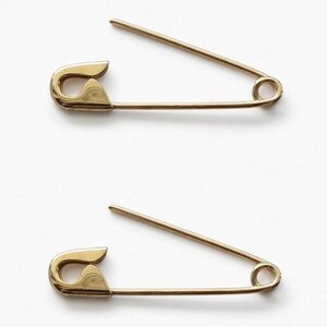 May include: Two gold-toned metal safety pins positioned horizontally. Each pin features a classic coiled spring mechanism at one end and a protective clasp at the other, with the sharp needle point currently open and angled upward from the main bar.