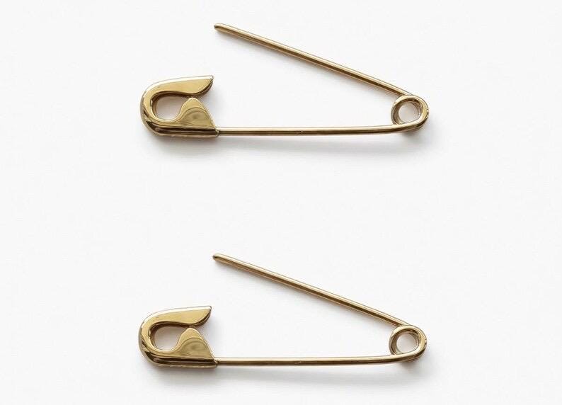 May include: Two gold-toned metal safety pins positioned horizontally. Each pin features a classic coiled spring mechanism at one end and a protective clasp at the other, with the sharp needle point currently open and angled upward from the main bar.
