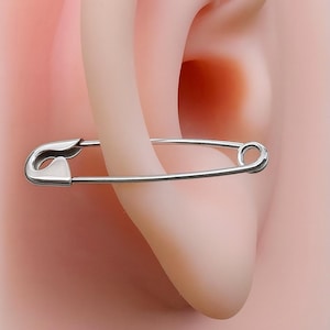 May include: A silver-toned safety pin earring featuring a polished metallic finish. The earring is designed to resemble a standard safety pin with a functional clasp mechanism on one end and a circular loop on the other, worn through the earlobe.