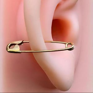May include: A gold-toned safety pin earring featuring a classic coil spring mechanism at one end and a pointed clasp at the other. The metal has a smooth, polished finish, and the design mimics the shape of a standard utility safety pin.