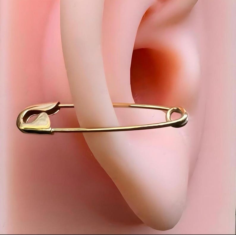 May include: A gold-toned safety pin earring featuring a classic coil spring mechanism at one end and a pointed clasp at the other. The metal has a smooth, polished finish, and the design mimics the shape of a standard utility safety pin.