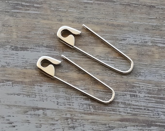 Könnte beinhalten: Ein Paar Ohrringe in Form von silberfarbenen Sicherheitsnadeln aus Metall. Jeder Ohrring verfügt über ein klassisches U-förmiges Drahtdesign mit einem funktionalen Verschlussmechanismus an der Oberseite und einer glatten, polierten Metalloberfläche.