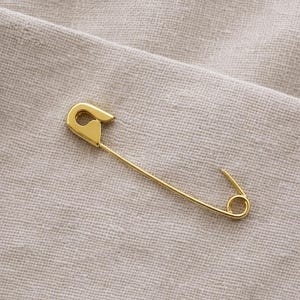 May include: A gold-coloured safety pin, positioned diagonally on a textured, light beige fabric. The pin features a classic design, with a curved top and a circular clasp. The pin's shiny surface reflects the light.