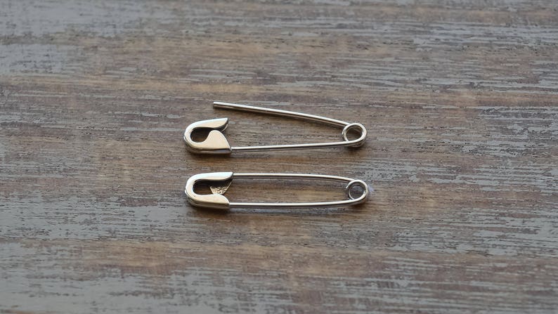 May include: Two silver-toned metal safety pins. The top safety pin is shown in an open position with the sharp needle point exposed, while the bottom safety pin is shown in a closed position with the needle secured inside the protective clasp.